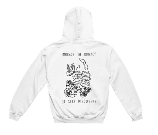 This white hoodie features a bold black-and-white graphic on the back, centered around a skeletal hand with roses growing around it and a butterfly resting on the fingers. The design is framed by the phrase “EMBRACE THE JOURNEY OF SELF DISCOVERY”